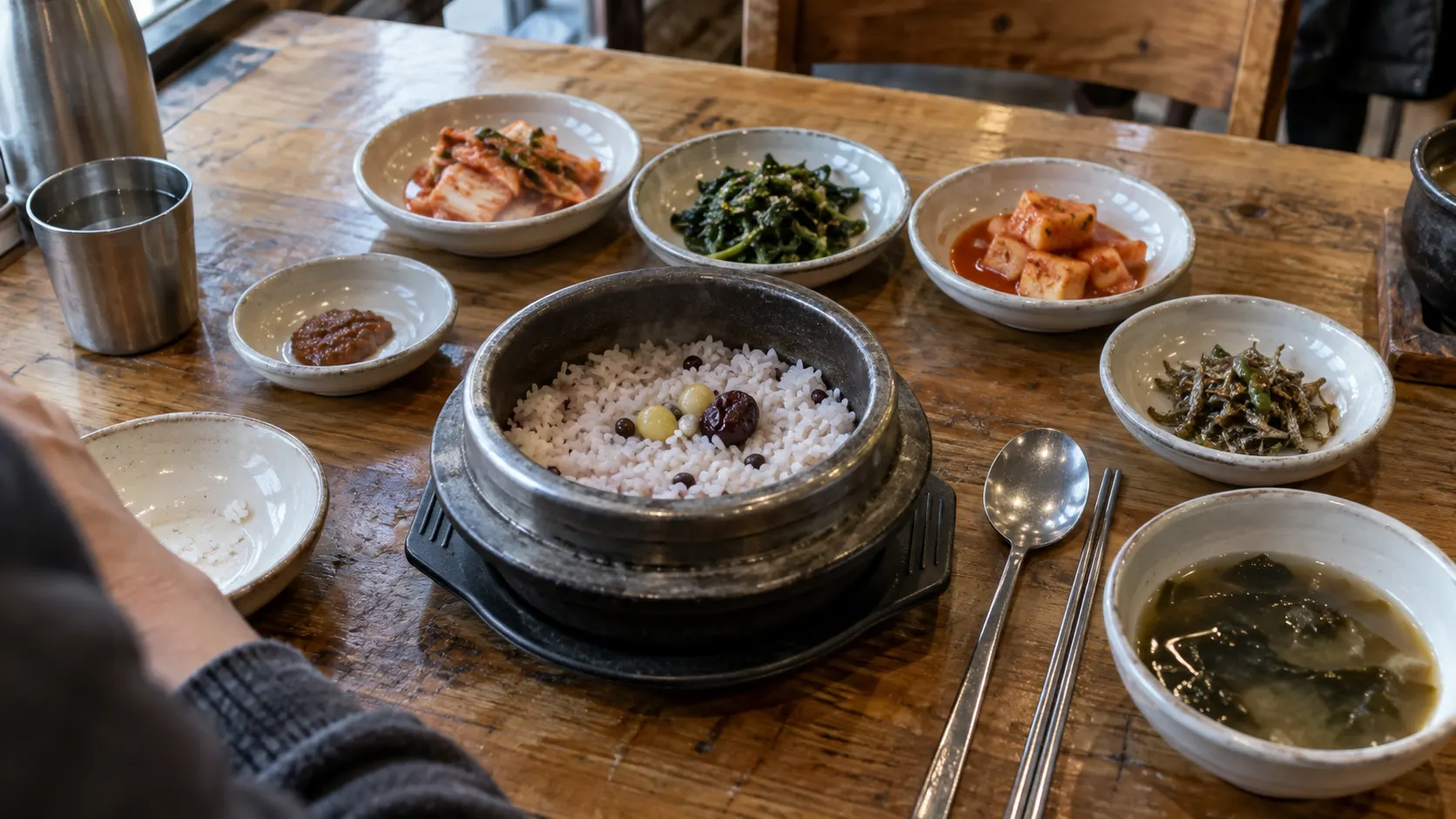 솥밥을 놓은 식탁에서 받침이 평평하게 닿는지 확인하는 장면입니다. 뜨거운 솥과 수저의 위치를 구분하고 주변 그릇의 간격을 살펴 손님이 식사를 시작하기 전 상차림 상태를 점검합니다.