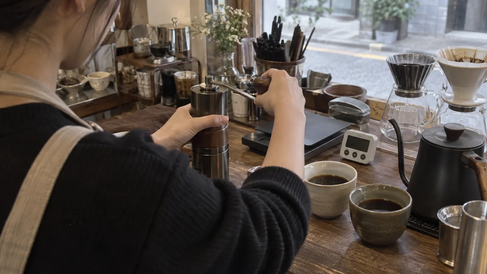 작은 카페 작업대에서 바리스타가 저울로 원두 투입량을 맞춘 뒤 분쇄도만 조절해 두 잔을 비교하는 모습입니다. 타이머와 추출량 기록으로 변수 점검 순서를 보여 줍니다.
