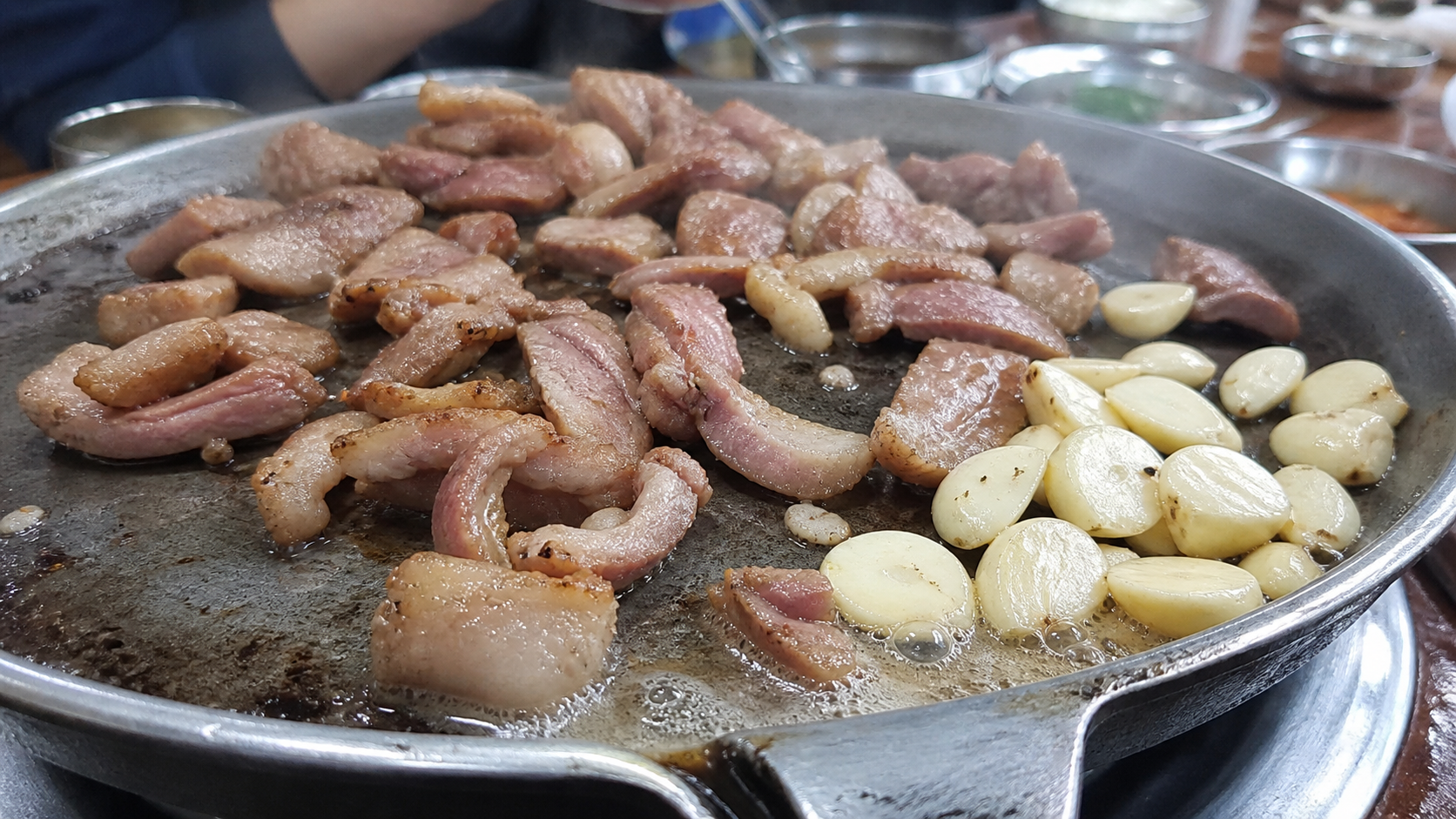 오리구이를 팬에 굽는 장면으로, 기름이 빠지는 과정과 마늘을 함께 올려 풍미를 더하는 조리 상황을 보여줍니다. — 오리구이의 기본 특징(오리구이에 대해 자주 묻는 질문과 알아둘 점) 내용을 보여주는 사진