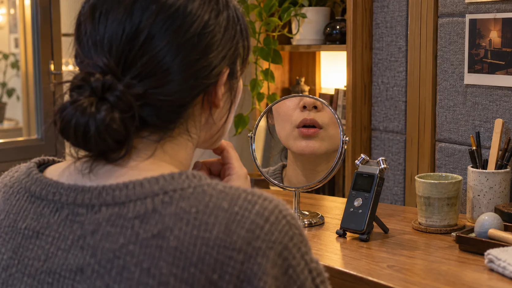 학습자가 작은 거울 앞에서 입술 모양을 살피며 모음을 말하는 장면입니다. 단어를 이루는 소리를 구별한 뒤 입 모양과 혀의 위치를 조절하고 녹음으로 원음과 비교하는 영어 발음 연습을 나타냅니다.