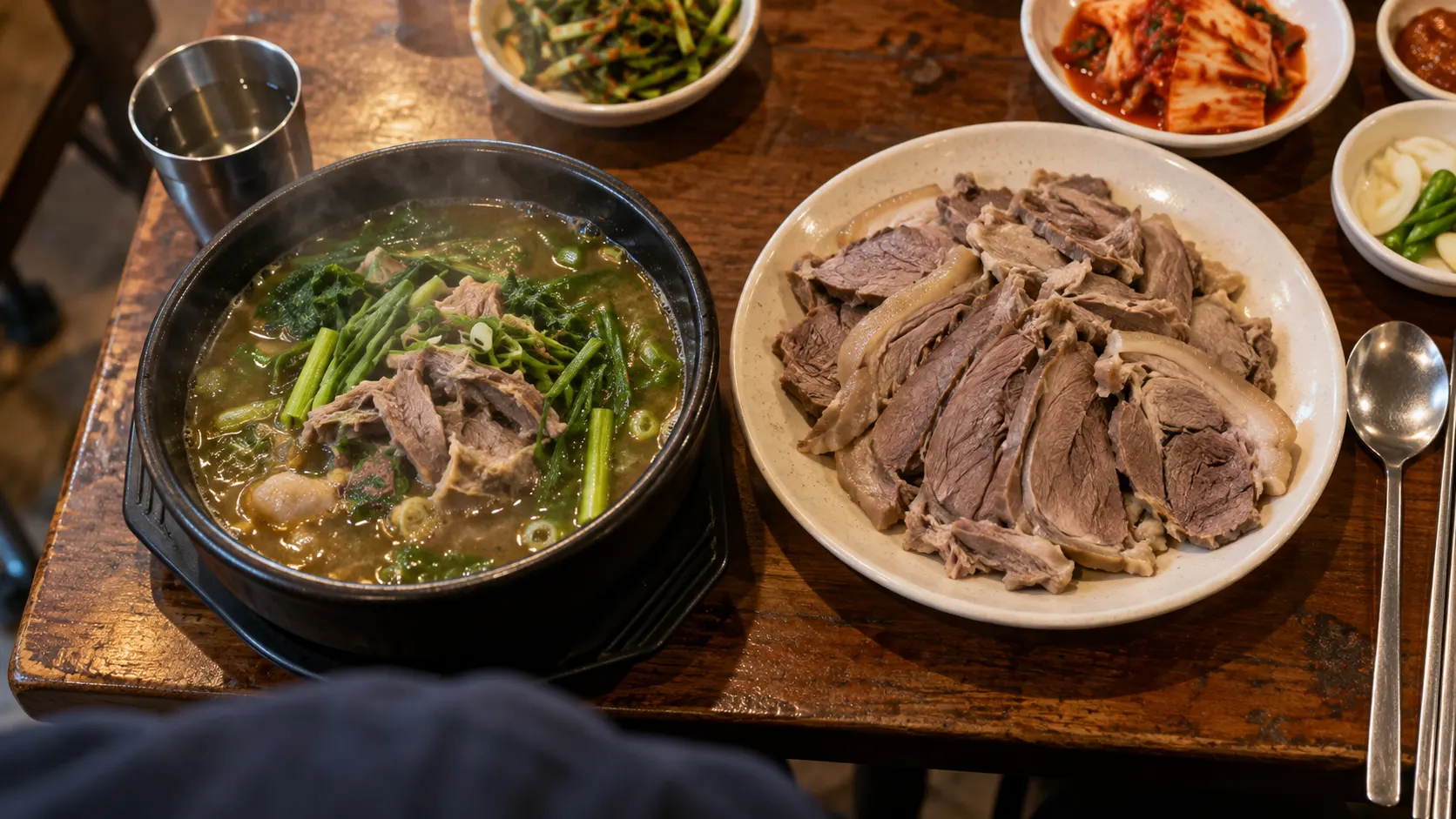 흑염소탕 한 그릇과 썰어 담은 수육 접시를 위에서 내려다본 모습입니다. 국물을 중심으로 먹는 형태와 고기를 따로 집어 먹는 형태가 나란히 놓여 식사 방식의 차이를 보여 줍니다.
