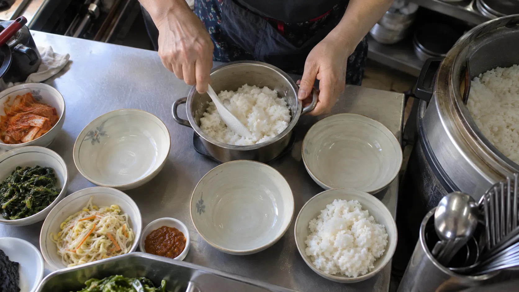 솥에 담은 밥과 곁들임 음식이 놓인 조리대에서 실제 제공 그릇에 음식을 담는 장면입니다. 촬영용으로 양을 늘리거나 기본 구성에 없는 반찬을 더했는지 대조하는 준비 과정을 보여 줍니다.