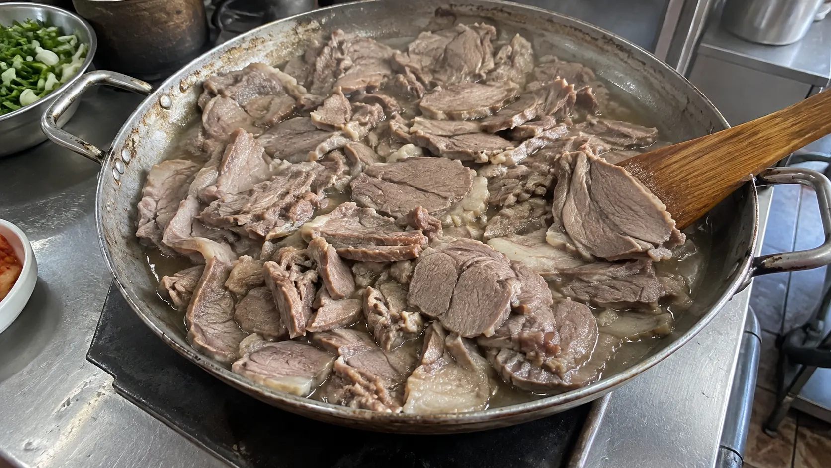 얕은 팬에 얇게 썬 흑염소 고기가 펼쳐져 있고 조각 사이에 조리 중 나온 수분이 고여 있습니다. 주걱으로 가장자리를 들어 고기 두께와 팬 바닥에 남은 액체를 함께 보여주는 장면입니다.