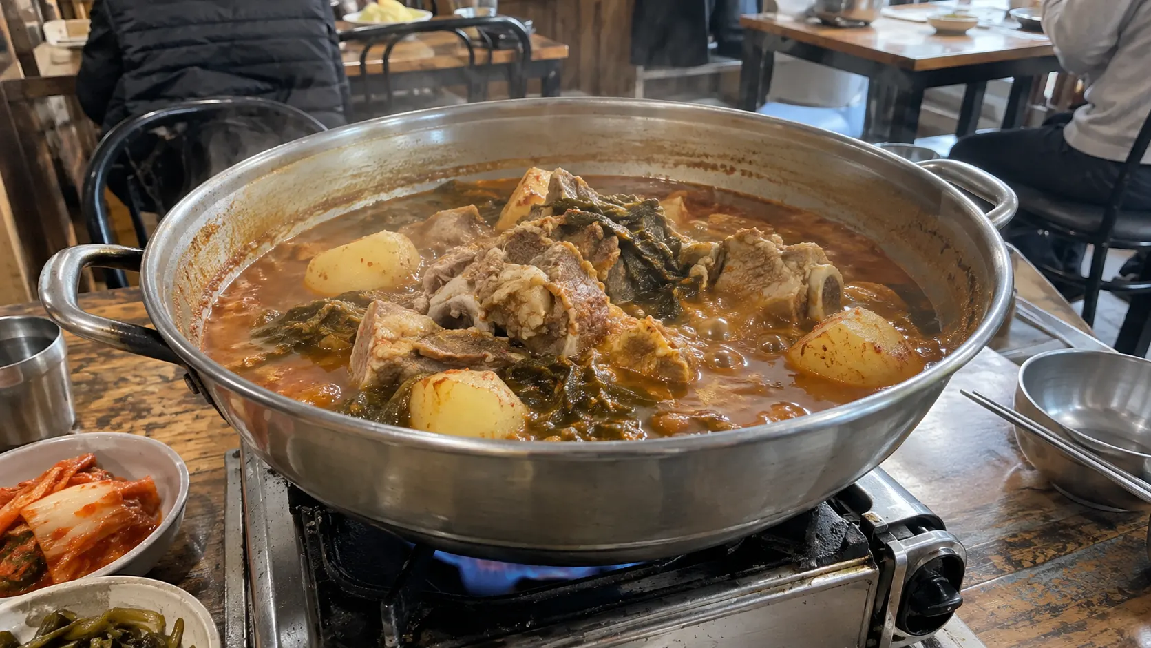 식탁 위 냄비에서 감자탕이 잔잔하게 끓고 있으며 냄비 안쪽에는 국물이 줄어든 흔적이 남아 있습니다. 국물 표면의 감자 조각과 액체의 높이를 함께 보여 주어 감자 풀림과 수분 감소를 구분하는 장면입니다.