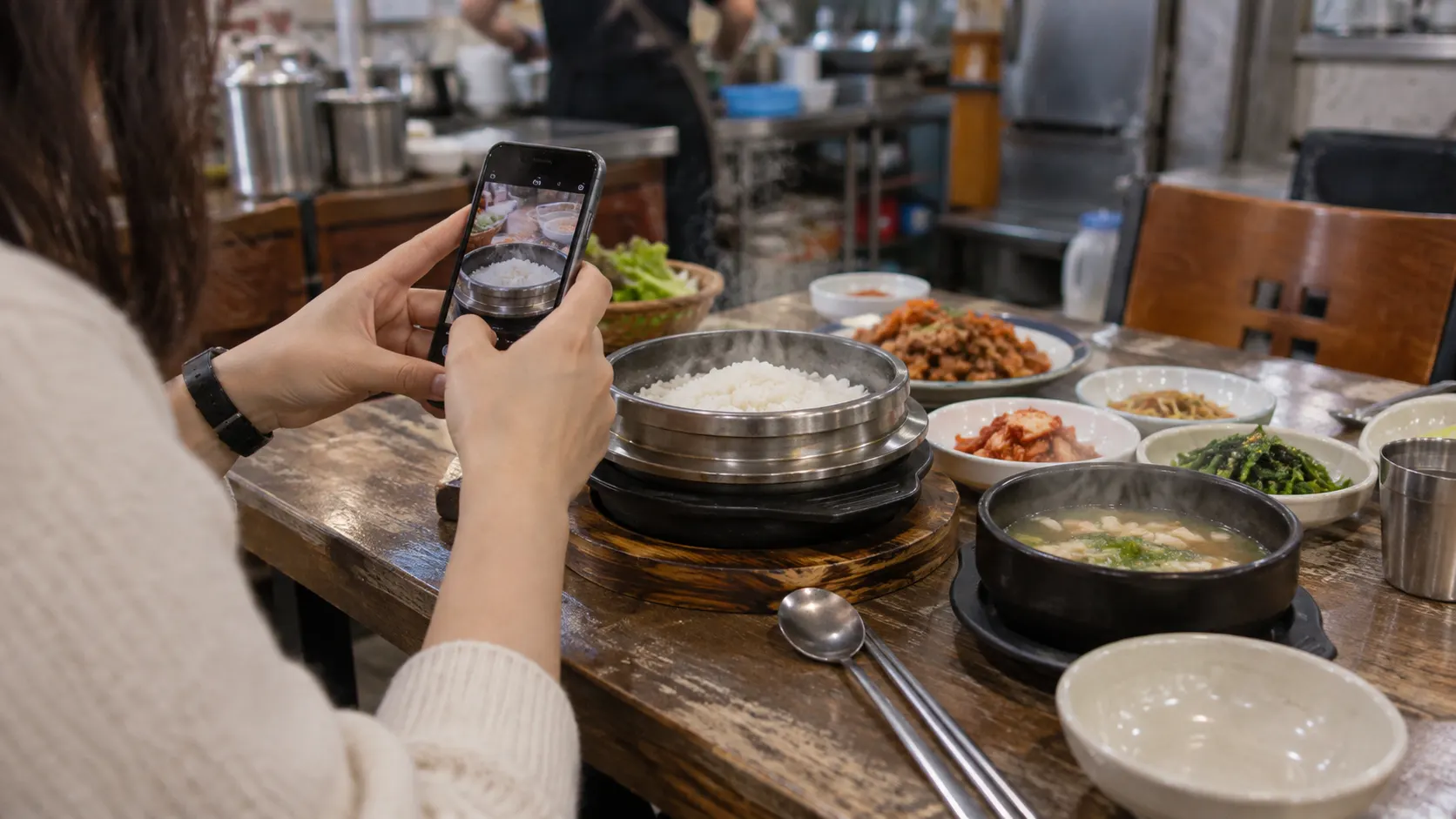 솥 가장자리 높이에서 휴대전화로 음식과 곁들임 접시를 촬영하는 장면입니다. 위에서 내려다볼 때 빠지는 그릇의 높이와 옆면을 담아 전체 상차림의 비율을 비교하는 구도를 보여 줍니다.