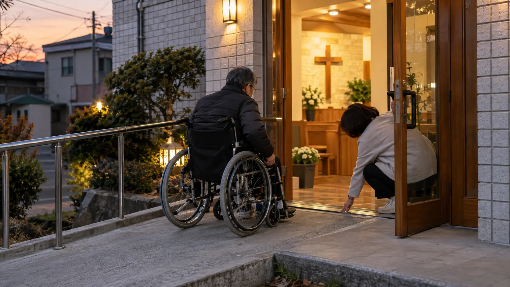 휠체어 이용자가 예배당 현관의 경사로를 지나고, 동행자가 문턱 높이와 문이 열리는 방향을 살피며 손잡이 주변의 대기 공간과 출입구 앞 대기 위치까지 확인하는 장면입니다.