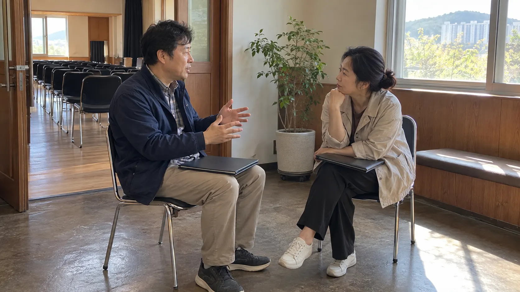 강의실 밖 휴식 공간에서 두 경영자가 마주 앉아 업무상 고민을 나누는 모습입니다. 동료의 판단 근거를 묻는 대화를 위해 강의 외 시간을 남기고 후속 논의의 주제를 정하는 상황입니다.