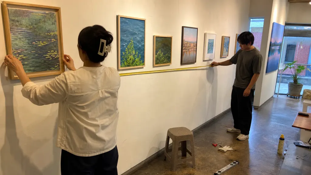 기획자와 작가가 전시장 벽면의 작품 간격을 줄자로 확인하며 주제에 맞는 배치와 관람 동선을 조정합니다. 작품마다 맡는 역할을 공간 안에서 구분하는 장면입니다.