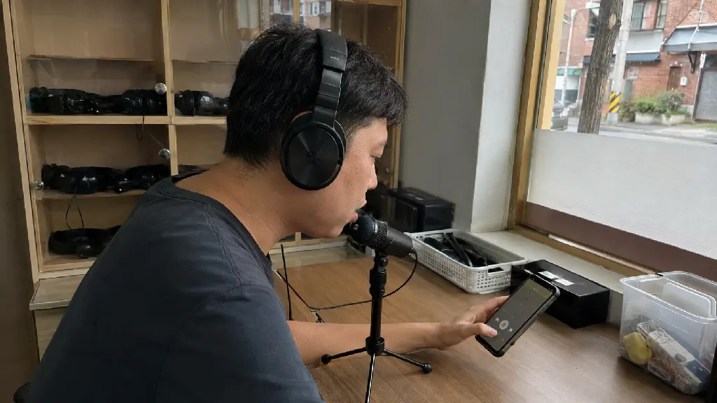 학습자가 헤드폰으로 짧은 영어 문장을 다시 듣고 스마트폰 화면을 확인한 뒤 입으로 따라 말하는 장면으로, 문장 단위 반복과 발화 전환의 순서를 보여줍니다.
