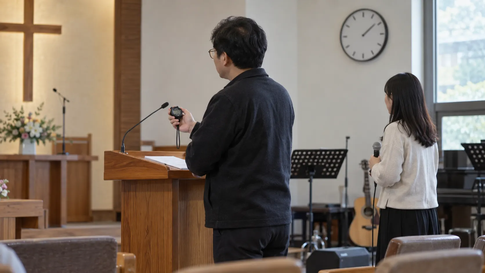 예배 진행자가 스톱워치로 찬양과 설교 사이의 전환 시간을 재고 벽시계와 담당자 위치를 기준으로 순서별 시작·종료 시각을 맞추는 장면입니다. 마이크 교체 시간도 별도 항목으로 계산합니다.