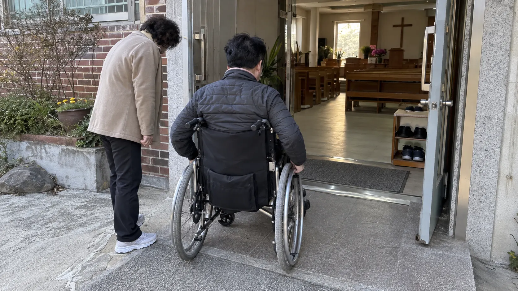 휠체어 사용자가 예배 공간 현관의 문턱과 열린 문을 지나며 회전할 자리와 안내 동선을 확인하는 장면입니다. 동행자도 옆에서 이동 폭을 살핍니다.
